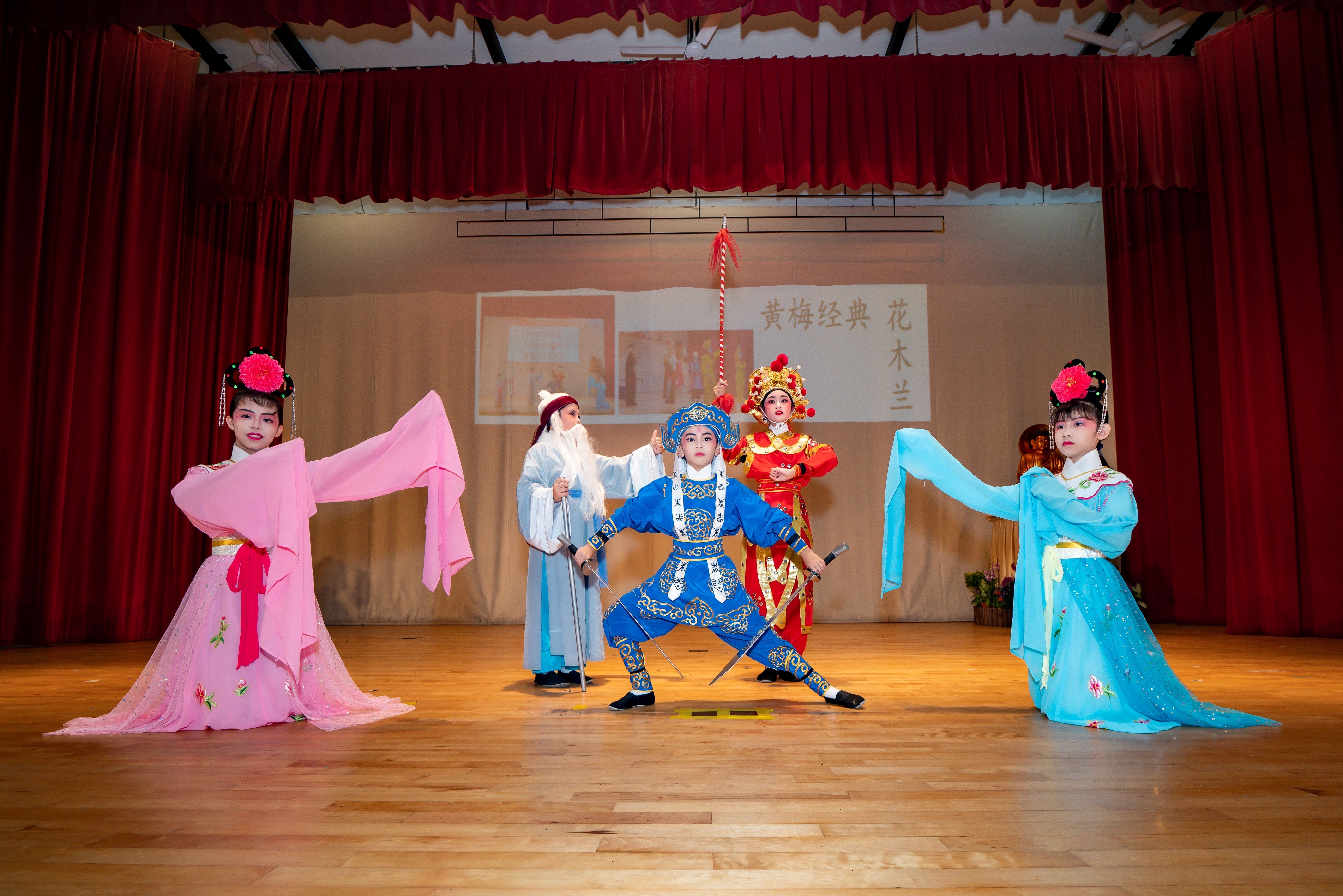 Five children in colorful traditional Chinese opera costumes perform on a wooden stage with red curtains.